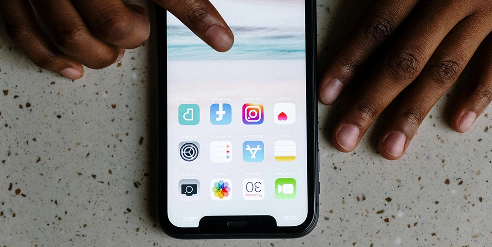 A hand taps on an iPhone screen, which displays various apps including Facebook, Instagram, and X, representing digital marketing services.